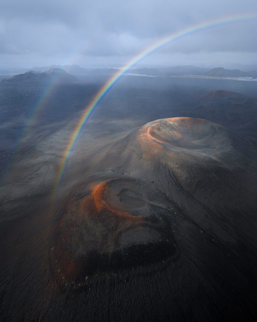 Aerial drone view of a rainbow arching over red cinder craters on a dark lava plain in the Icelandic Highlands.