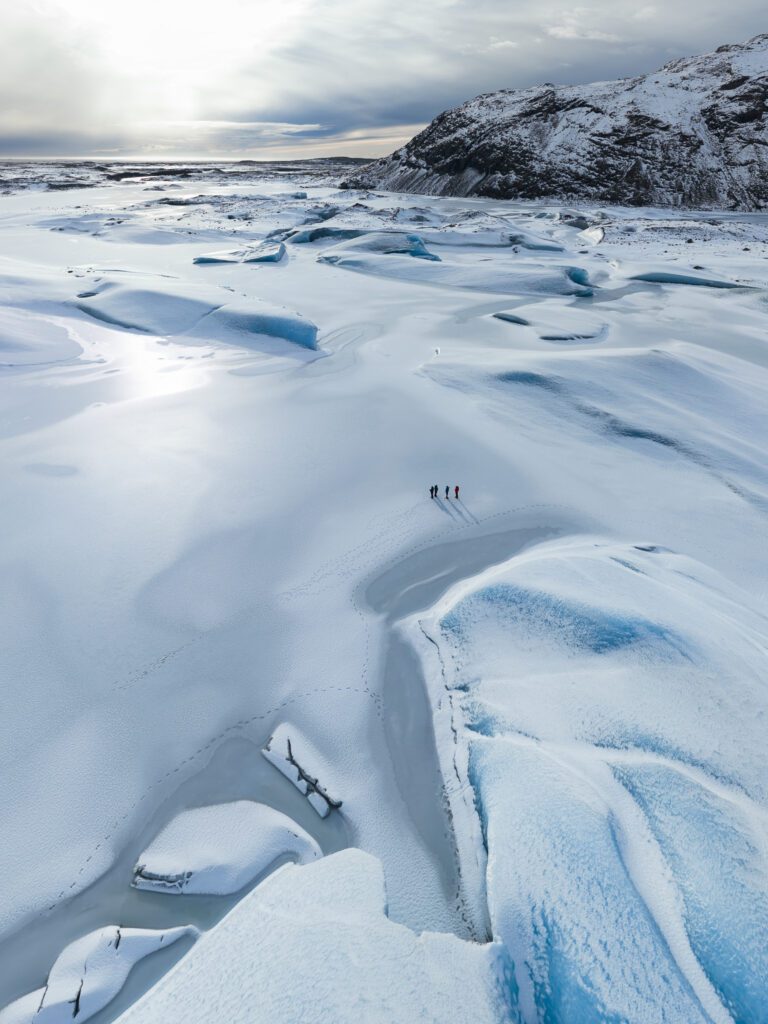 Tiny figures crossing a snow-covered glacier tongue beneath a dark mountain ridge in south Iceland.