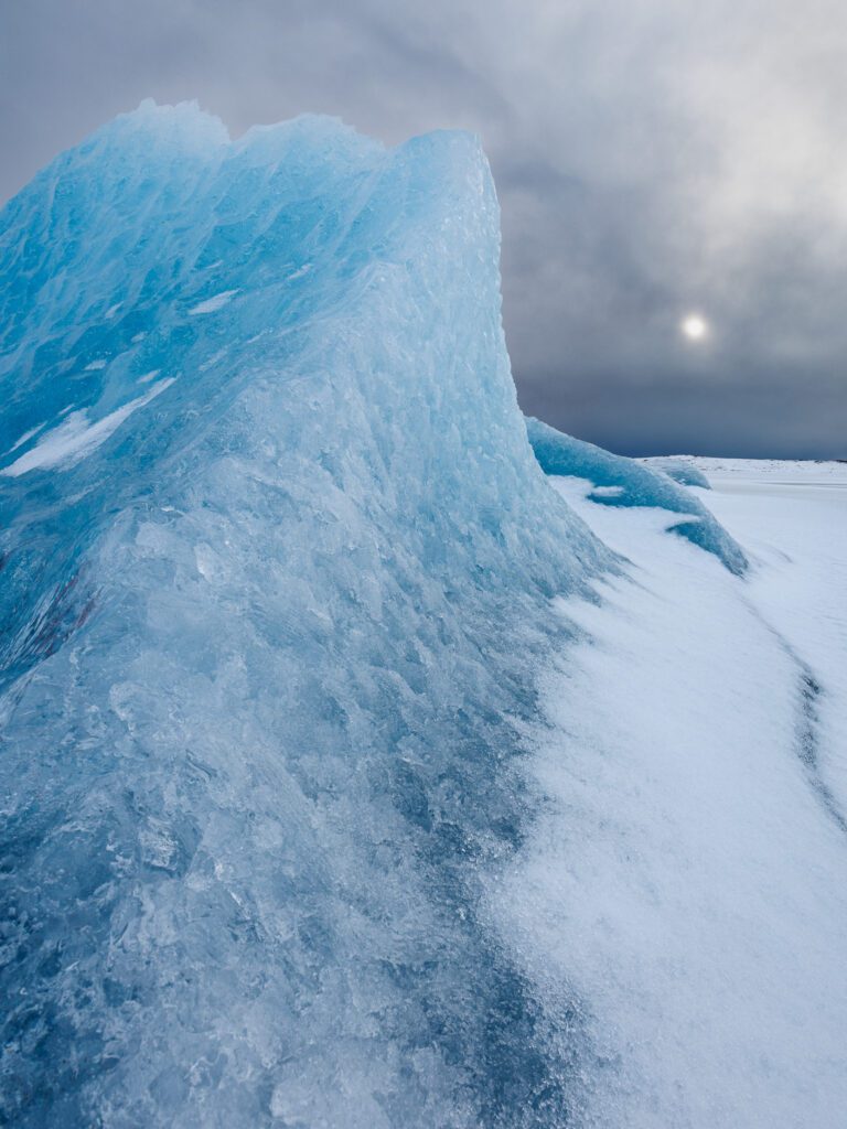 Sculpted pale blue iceberg rising from a snowfield with a dim sun behind grey cloud.